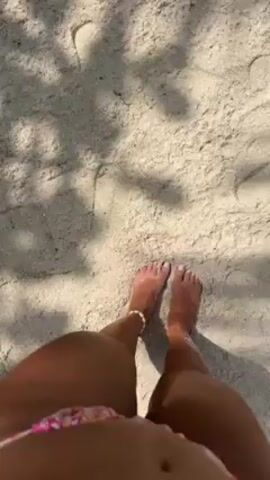 The video begins with a view of a person's bare feet standing on a sandy surface. The feet have a light tan and are adorned with a shell anklet on the left ankle. The camera angle looks down on the person's legs, revealing a portion of their floral patterned bikini. The sandy ground is uneven and marked with footprints. The shadow of foliage is cast over the sand. As the camera moves, the view expands to show the sandy beach, and then pans upwards to reveal palm trees and the turquoise ocean in the background. People are lounging on the beach under an umbrella. The scene suggests a tropical beach setting with white sand, clear water, and swaying palm trees.