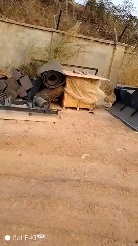 The video depicts an outdoor scene with various metal components and structures scattered on a dirt surface. There's a concrete wall with barbed wire along the top and tall grass in the background. The metal components include what appears to be parts of machinery or equipment, some stacked and some lying flat on the ground. A wooden crate is also visible. The video pans across the scene, showing the arrangement of these objects in relation to the wall and the surrounding environment. The lighting is somewhat overcast, and the overall impression is of an industrial or storage area. There are no people visible in the video.
