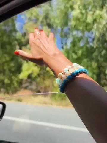 A person's arm with bracelets is extended out of a car window. The hand is waving. The background is a blurred landscape with trees and blue sky.