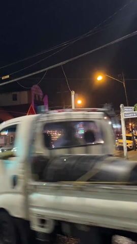 The scene is a nighttime shot of a street intersection. Cars are stopped at the intersection, waiting for the light to change. A truck with flashing lights is in the foreground. A street lamp illuminates the scene. The background features buildings with signage and lighting. Some electrical wires crisscross above the street.