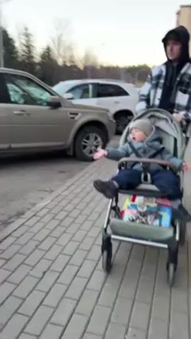 A young man pushes a toddler in a stroller along a paved sidewalk next to a parking lot. The toddler is wearing a winter hat, jacket, and boots. The man is wearing a black hooded sweatshirt, and a black and white plaid shirt. There are cars parked in the lot to the left. The toddler gesticulates and appears to be talking.