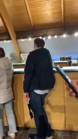 The video shows a group of people inside a building with a distinctive arched wooden ceiling. The people are gathered around a long counter, possibly a bar or a tasting area. Most of them are standing and facing the counter, likely waiting for service or tasting something. Some are wearing winter coats, suggesting it's a cool environment. The woman in the foreground is wearing glasses and a dark jacket and is holding a glass, possibly of wine. The building appears to be well-lit and has a rustic, perhaps wine-related, ambiance.