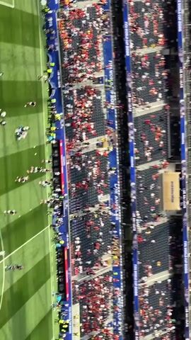 The video shows an aerial view of a soccer field after a game. A large number of people, likely players and staff, are scattered across the field, some lying down, others standing or walking. Confetti is visible on the grass. Spectators are seated in the stands surrounding the field. The scene appears to be a celebration or post-game ceremony.
