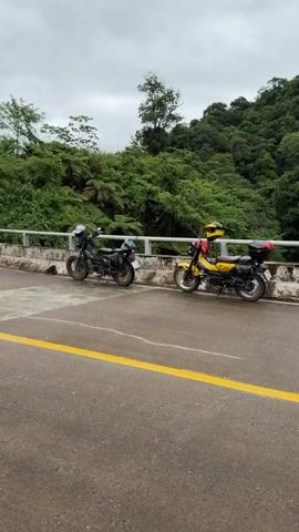 A POV shot shows a woman with blonde hair wearing a hooded garment and a backpack, standing on a road with two motorcycles parked nearby. The road curves uphill, surrounded by lush green vegetation and trees. The sky is overcast, suggesting a cloudy day.