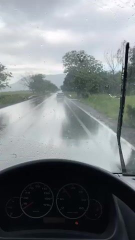 The video shows a first-person view from inside a vehicle driving on a wet road during a rainy day. Raindrops cover the windshield, partially obstructing the view. Windshield wipers are in motion, clearing some of the water. The dashboard and steering wheel are visible. The speedometer reads about 40 kilometers per hour. Outside, the road is flanked by green fields and trees. Another vehicle is visible further down the road. The sky is overcast with grey clouds.
