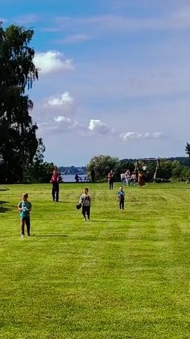 A group of children are running across a grassy field on a sunny day with a blue sky and white clouds. In the background, there is a body of water and a tree line. There are also a few adults standing in the background, some of whom appear to be watching the children. One of the children in the foreground is holding a blue water bottle.