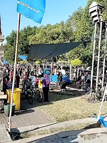 A daytime outdoor video captures various scenes in what appears to be a public park or square. The video begins with a shot of several people gathered under a black tarp canopy, with a blue flag visible on the left side of the frame. There's also a tall structure resembling a birdhouse on a pole to the right. The next shot shows multiple white flags with blue logos and a cartoon robot character, likely advertisements, flapping in the wind. A subsequent scene features a large monument with a blank electronic billboard and a water feature with several people sitting along its edge. The video concludes with a close-up of a large fountain with water shooting high into the air, surrounded by trees and buildings in the background. Throughout the video, the sky is clear and blue, suggesting good weather.