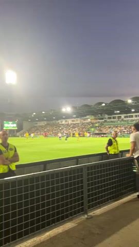 The video captures a football match in a stadium under a twilight sky. The scene is set in Žilina, Slovakia, as indicated. The stadium is illuminated by bright floodlights, casting a glow on the lush green pitch where players in yellow and red jerseys are engaged in a game. Spectators fill the stands, their faces turned towards the action. In the foreground, a metal fence with wire mesh separates the spectators from the pitch. Two individuals in yellow high-visibility vests, likely security personnel, stand guard near the fence. A man in a white t-shirt and black shorts leans against the fence, watching the game. The sky is a deep, dusky blue, suggesting the time of day is evening, possibly late dusk. The overall atmosphere appears to be one of enthusiastic engagement with the sporting event. Visible text on a scoreboard in the distance shows "0:0", indicating the current score.