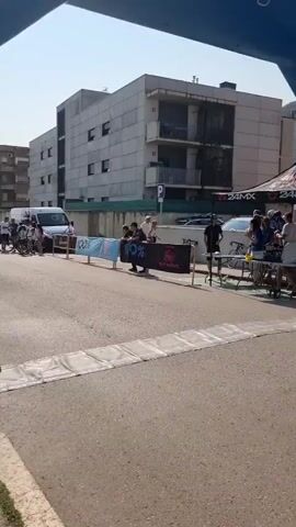 The video shows a street scene with a blue inflatable archway at the foreground, likely marking the start or finish line of a race. Several people are gathered under tents behind a barrier. One person in a red cycling jersey and white helmet is riding a bicycle towards the archway. Another cyclist is visible further in the background. Buildings and tents are situated on the sides of the street. The weather appears sunny.
