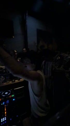 The video depicts a nightclub or bar scene. A DJ is performing, visible behind a DJ console with illuminated buttons and dials. In front of the console, a person wearing a white tank top and camouflage pants is dancing energetically with their arms raised, seemingly enjoying the music. The lighting is dim, with only a few light sources illuminating the area. A small crowd of people is visible in the background, mostly standing and watching. Some people are seated at the bar to the side. The environment appears to be a social gathering centered around music and dance in a dimly lit indoor venue.