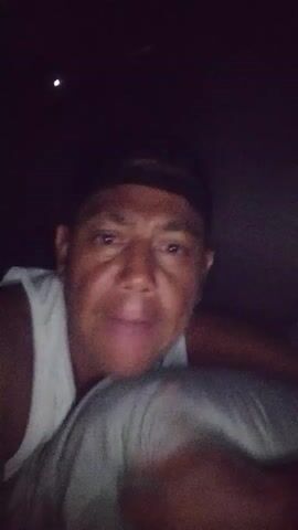 A dark video shows a close-up of a person's face. The person appears to be lying down and holding a pillow or blanket. The person is wearing a white tank top and a black baseball cap. The person's eyes are open and they are looking directly at the camera. Their mouth is slightly open as if speaking. The background is very dark, with a single small light source visible at the top left of the frame. The person's facial expression is difficult to interpret due to the low light and close-up perspective. The video appears to be a selfie or a recording from a handheld device.