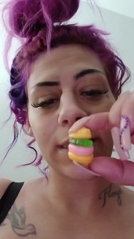 Here is a description of the video:
The video shows a woman holding a small, colorful gummy burger between her fingers. The burger has layers of yellow, pink, and green, resembling the bun, patty, and lettuce. The woman has light skin, long dark eyelashes, and purple and dark hair styled in an updo with curly strands falling down. She has a tattoo of a bird on her chest and the word "Floyd" written near her collarbone. Her fingernails are long and manicured, with white polish and purple accents. She looks directly at the camera with a slightly pursed expression. The background is a plain, off-white wall.
