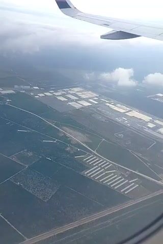 From the window of a Volaris airplane, the landscape below is visible. Large rectangular structures, possibly greenhouses or warehouses, dominate the view alongside fields and a long series of structures that could be poultry houses. A road cuts across the lower portion of the frame. The sky is partly cloudy.