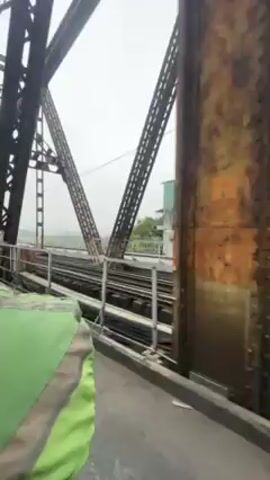 The camera, seemingly mounted on a vehicle, travels across a metal bridge. A complex network of rusted metal beams and trusses forms the bridge's superstructure, visible on the left.  To the right of the camera's path, train tracks run parallel to the road.  In the distance, on the bridge ahead, a couple of individuals are visible standing near the tracks. The sky is overcast, creating a muted color palette for the scene.  A section of a green backpack or bag is visible in the bottom left corner of the frame, suggesting the viewpoint is from someone wearing or carrying it. The video's setting appears to be a bridge with mixed transportation infrastructure, combining rail and road over a wide, pale landscape.