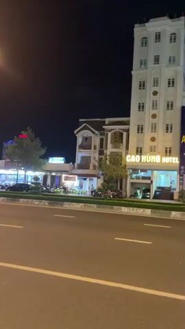 At night, a streetscape in Vietnam is visible, with a focus on a brightly lit area across the street. The camera initially focuses on a tall white building with multiple windows, identified as Cao Hùng Hotel by the sign on its facade. Adjacent to the hotel is a large sign advertising An Dương Phi, associated with CTy TNHH. The camera slowly pans right to reveal a strip of businesses and restaurants, heavily illuminated with bright lights and decorations. Many motorcycles are parked in front of these establishments. Streetlights cast a warm glow on the road, which is lined with a median strip of low greenery. Few cars or pedestrians are visible. The sky is dark, indicating the late hour.