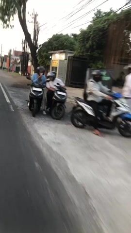 The video shows a street scene with several motorcycles and people. In the foreground, the road is visible with a white line marking the edge of the lane. The scene is slightly blurry, suggesting movement. On the side of the road, two people on a motorcycle are stopped. The rider is wearing a blue shirt and a helmet, and the passenger is wearing a white shirt and a helmet. Further down the road, another motorcycle with two people is in motion, also wearing helmets. The rider appears to be wearing a light-colored shirt and a dark mask, and the passenger is in a light-colored jacket. In the background, there are buildings, trees, and power lines. The overall impression is a snapshot of everyday life on a street, possibly in a warm climate given the clothing.