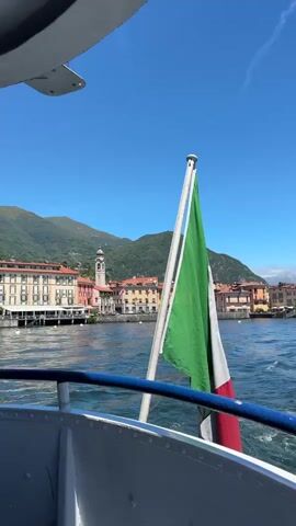 The video begins with a close-up of an Italian flag waving on a boat as it moves across a body of water. In the background, a picturesque town with colorful buildings and a church steeple is visible on the shore, nestled against a backdrop of lush green mountains under a clear blue sky. The camera angle suggests the viewer is on the deck of the boat, looking out towards the town. The water is choppy, indicating the boat is in motion. The scene evokes a sense of travel and scenic beauty, likely in Italy.