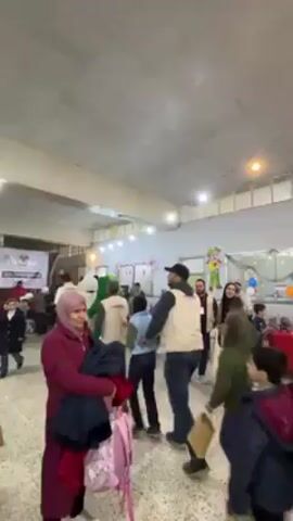 The video shows an indoor event filled with people, mostly children and adults, in a large room. The room appears to be a hall or community center, indicated by the white ceiling, fluorescent lighting, and the presence of decorations, including streamers and a large backdrop with text in Arabic. On stage, there is a man in a white suit, a person dressed in a panda costume, and another in a green costume, possibly for entertainment. People are walking around, some interacting with each other. The atmosphere seems festive and lively. A Syrian flag is visible in the background.