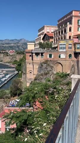 The video shows a high-angle view of a coastal town with buildings built on cliffs overlooking a harbor filled with boats. The buildings are a mix of colors, including pink, peach, and tan. Many of the buildings have multiple stories and balconies. The harbor has a long pier with many small white boats docked. The water is blue, and the sky is clear and blue. Mountains are visible in the background. Lush green trees and plants are in the foreground, obscuring some of the lower buildings. Some people are visible on a terrace area with tables and umbrellas. Cars are parked on a street below the buildings.