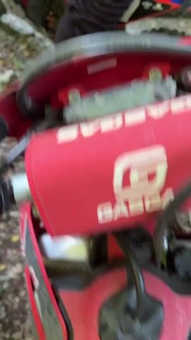 The video shows a close-up of a red and white dirt bike, with the "GASGAS" logo visible on the handlebars. The bike is parked on a dirt path surrounded by trees and foliage. The rider's legs, clad in black pants and red and white boots, are visible. The camera then pans to show the rear tire of the motorcycle on a muddy, leaf-covered path, before moving forward into a wooded area with dappled sunlight filtering through the trees. The footage is shaky, suggesting it was recorded from the perspective of someone riding or walking behind the motorcycle.