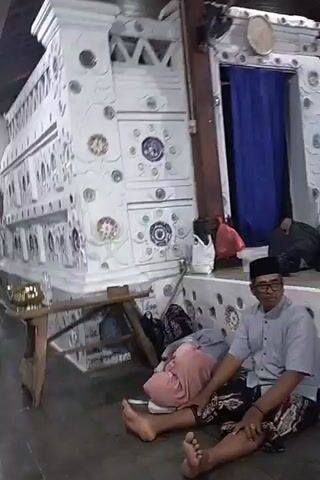 The video shows an interior scene with people sitting on the floor and on a raised platform. The walls are decorated with white tiles and circular patterns. In the foreground, a man wearing a light blue shirt, glasses, and a black cap sits on the floor, facing forward. To his left, another person is lying down, partially covered by a pink cloth. In the background, several people are seated on a raised platform. One woman wears a blue headscarf, and another wears a dark headscarf. A blue curtain hangs behind them. The space appears to be a covered outdoor area or a room with an open roof structure supported by wooden beams. Various objects, including bags and what appear to be grave markers, are visible in the background.