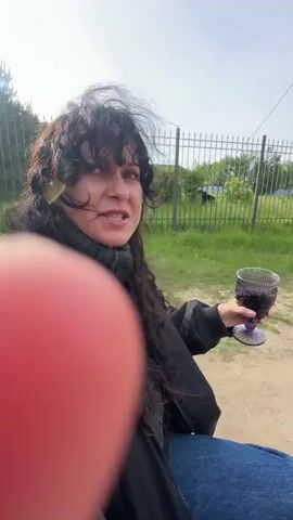 The video shows a woman with long, dark, curly hair and a black jacket. She is holding a decorative purple glass filled with a dark liquid. She is outdoors, with a fence, trees, and a bright sky in the background. Her hair is being blown by the wind. The woman smiles and turns her head to look at the camera. The video is shot from a close-up perspective, with a blurred object, possibly a finger, in the foreground.