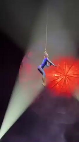 A stage aerialist, seen from the back, hangs in the air by a rope attached to her hair. She wears a blue jumpsuit. She is illuminated by spotlights, two white and one red, with theatrical smoke swirling around her. Behind her is a large, round prop colored red. Spectators sit in the foreground. She slowly rotates and poses.