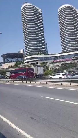 The video shows a daytime view from a moving vehicle on a highway. In the background, several tall, modern skyscrapers with curved, white facades dominate the skyline. Below the skyscrapers, a large building complex with a glass facade and a flat roof is visible, possibly a mall or commercial center. Various vehicles, including cars, a white van, a red and white truck, and a white trailer truck, are driving on the highway. Some of the vehicles are driving in the same direction as the camera, while others are going in the opposite direction. The highway has multiple lanes and is divided by a barrier. The sky is clear and blue, indicating sunny weather.