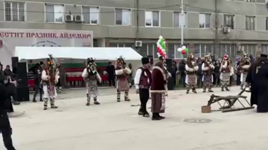 The video shows a group of people, some wearing traditional Bulgarian Kukeri masks and costumes, performing a ritual in an outdoor public space. They are gathered around a wooden plow or sleigh. One individual, dressed in priestly garb, stands to the right of the plow. A few other people are dressed in traditional clothing. A banner in Bulgarian is visible in the background, and a white tent can be seen on the left. The setting appears to be a town square in front of a multi-story building. A crowd of onlookers stands behind the performers, and Bulgarian flags are prominently displayed. A traditional folk tune is playing.