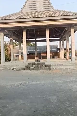 The video begins with a view of a building under construction, possibly a pavilion or a gazebo, with a tiled roof supported by concrete pillars. In the foreground, a stack of concrete blocks is visible. The camera then pans to the right, showing a dark-colored motorcycle parked on a paved surface. The motorcycle has a license plate with the text "S 6569 BAA". A person, partially visible, is sitting on the motorcycle. The camera continues to move, focusing on a ramp and stairs leading up to an area with beige tiles. The ground appears to be a mixture of concrete and gravel. The scene is outdoors, with vegetation and buildings visible in the background. The lighting suggests it is daytime, possibly late afternoon.