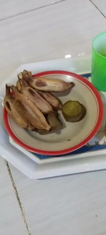A serving tray is shown from a top-down perspective. On the tray is a plate with several cooked bananas and a small piece of sweet potato. Next to the plate are four cups, three of which contain beverages: one with a light brown liquid, another with clear liquid, and a third with a dark brown liquid. The fourth cup is empty and green. A pink lid sits on top of one of the cups. The tray rests on a tiled surface.