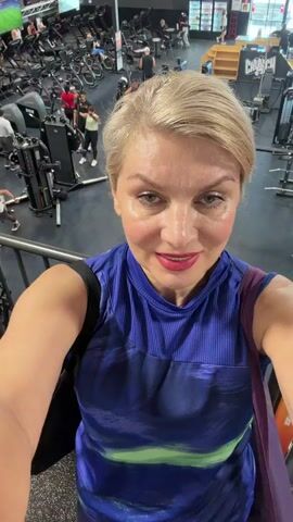A woman with blonde hair and makeup smiles at the camera as she stands on a walkway overlooking a busy gym. She is wearing a blue athletic top with a multi-colored design. Her arms are visible, and she is holding bags over her shoulders. The gym below is filled with people exercising on various machines, including treadmills, weight machines, and exercise bikes. The environment appears to be a modern gym with metal structures and equipment. The lighting is bright, suggesting it is daytime.