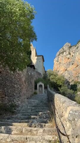 A stone staircase ascends towards a building with a bell tower. A wall made of rough-hewn stones runs along the left side of the staircase. To the right of the staircase, there is a stone railing. The staircase leads to a small shrine-like structure with a pitched roof, just before reaching the building. Above the building, a rocky cliff face rises, covered in sparse vegetation. A large tree with green leaves is visible on the left side of the frame. The sky is a clear, vibrant blue. The sun appears to be shining, casting shadows on the stairs and walls. The scene is an outdoor view of an ancient or historical architectural setting in a natural environment. There are no people or animals visible in the video. The camera is stationary, capturing a still image of the scene.