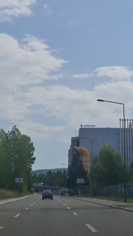 The video shows a view from a moving vehicle on a road. The road has two lanes and is paved. On the right side of the road, there is a fence and trees. Further in the background, there is a large building with the "swisscom" logo visible. There are also several large yellow pipes extending from the side of the building. On the left side of the road, there are trees and a road sign. The sky is blue with scattered clouds. A few cars are visible on the road, including the vehicle from which the video is being recorded.