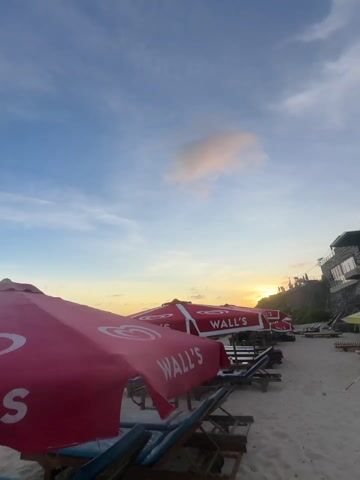 The video starts with a low-angle shot of beach umbrellas branded with the "Wall's" logo, silhouetted against a sunset sky. The camera then pans down to reveal a woman in a blue bikini, wearing sunglasses and a yellow wristband. She appears to be taking a selfie. The camera continues to angle upwards, focusing on the sky with the woman's silhouette against it, as she looks up. The scene is a sandy beach with a blue sky, a few clouds, and beach umbrellas.