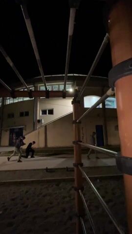 The video captures a nighttime scene, looking out from underneath a playground structure towards a building. In the immediate foreground, dark, granular ground, likely sand or wood chips, is visible. Above this, a network of vertical brown poles and horizontal silver bars crisscrosses, forming part of a climbing frame or monkey bars. The camera is positioned low, looking up through these bars.

In the mid-ground, there is a paved pathway or courtyard. On this path, two individuals are engaged in physical activity. One person, wearing a light-colored long-sleeved top and dark pants, is performing lunges. They step forward with one leg, bending both knees, then push back up. Near them, another person, dressed in dark clothing, is crouched low to the ground, seemingly observing or assisting. Small, dark objects resembling dumbbells or weights are visible on the ground beside them. Another person in a dark jacket is seen walking from left to right further down the path.

In the background, a large, light-colored building stands. It has a curved or vaulted roofline and several windows, some of which are illuminated from within, casting a soft glow. There are also darker door openings visible. A bright light fixture on the building's exterior wall illuminates the area directly in front of it. Further to the right, another indistinct figure is partially visible near the building. The overall atmosphere is dark, with artificial lights providing the primary illumination for the scene, suggesting an evening or night workout session.