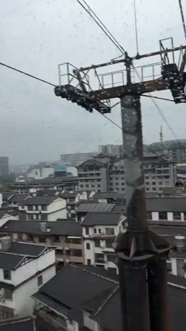 Here's a description of the video:

The video shows a view from inside a moving cable car, offering glimpses of the surrounding urban landscape on a cloudy day.

The camera is positioned inside the red-framed cable car, looking out through the window which is covered in raindrops. The view shows a dense cityscape below, composed of primarily white buildings with dark roofs. Tall buildings are visible in the background. A cable and support structure of the cable car system are also visible through the window.

Inside the cable car, several people are present. One person is taking photos with a mobile phone. Other people are looking out the window, observing the scenery.

The video seems to have been taken during a ride on the cable car, capturing the journey and the surrounding views.