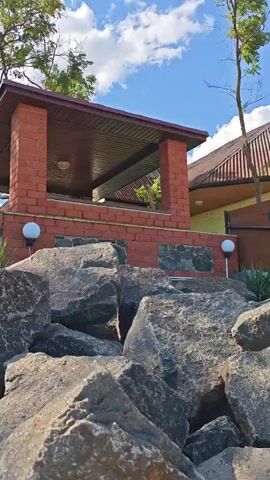 This is a video that shows a house with a large stone embankment in front of it. The house is yellow with a brown roof and has a porch. There are trees around the house and the sky is blue with some clouds. The video is taken from a low angle, looking up at the house.