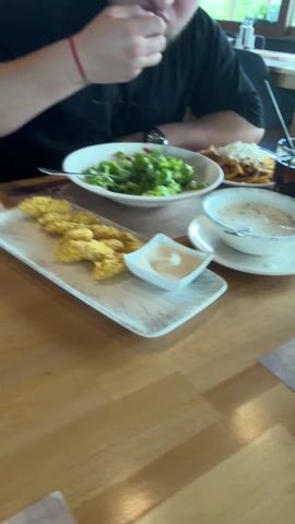 The video shows a person at a restaurant table. In front of them is a plate of crispy chicken strips, a bowl of creamy dipping sauce, a salad, and a bowl of soup. The person dips a chicken strip into the sauce. The table is wooden. A second person is visible in the background.