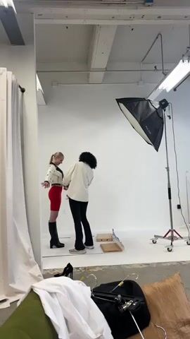 A blonde woman in a white jacket with black accents and red shorts stands in front of a white backdrop in a studio. She is being adjusted by a person with dark curly hair wearing a cream-colored top and black pants. The person is adjusting the blonde woman's jacket. In the foreground, there are clothes and cushions. The studio has professional lighting equipment, including a large softbox on a stand.