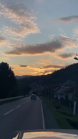 The video shows a view from a vehicle driving on a road towards a village nestled in a valley at sunset. The sky is filled with scattered clouds tinged with orange and yellow hues from the setting sun. The road ahead curves gently, and a blue car is visible in the distance. On the right side of the road, houses with dark roofs line the hillside, and trees flank the left side of the road. Power lines and a utility pole are also visible. The reflection of the sunset can be seen on the vehicle's dashboard in the foreground. The overall scene depicts a peaceful, rural landscape during a picturesque sunset.