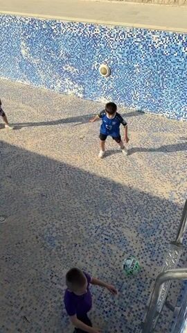 Several children are in an empty swimming pool, playing with a soccer ball. The pool is lined with small blue and white tiles, and has a ladder at one end. One boy wears a blue soccer uniform, another wears a white tank top and dark shorts, and a third is in a purple shirt. They kick and chase the ball across the tiled surface. The video is taken from a high angle, looking down into the pool.