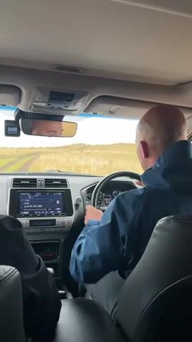 The interior of a car is shown, with a man in a blue jacket driving on a dirt road through a field of dry grass. The man is bald and his head is turned towards the road ahead. The car's dashboard, including a radio and climate controls, is visible. A rearview mirror reflects the face of another person in the back seat. The interior of the car is otherwise dark, with black leather seats. The scene is taken from the passenger seat, looking towards the driver. The road in front of the car is rough and uneven.