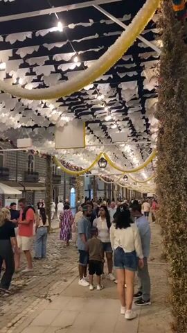 The video shows a crowded cobblestone street at night, decorated with white and yellow streamers and lights. People are walking in both directions, some in groups and some individually. Buildings with arched windows are visible on the left side of the street. The overall atmosphere appears festive, possibly during a street fair or festival.
