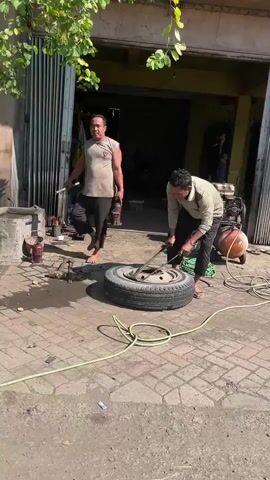 Two men are working on a car tire outside a garage. The man on the right is using a tool to pry the tire from the rim. The man on the left is standing and holding a metal bar. A spare tire, a compressor, and various tools are visible. The ground is paved with brick. The area appears to be an outdoor workshop or repair shop.