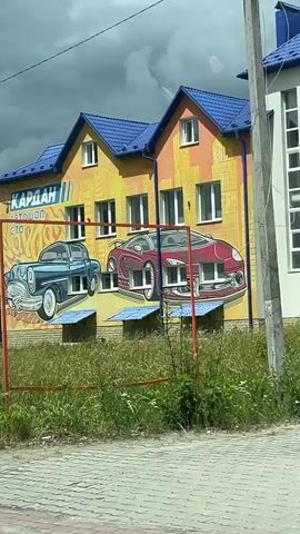 The video shows a passing view of a building with murals of cars on its exterior. The building is yellow with a blue roof and appears to be a business related to cars, indicated by the text on the building. There is a sign that says "РЕМОНТ СТАРТЕРА ГЕНЕРАТОРА, ТУРБИН", which translates to "REPAIR OF STARTER, GENERATOR, TURBINE". There are also stacks of tires visible. The environment is outdoors with green grass and trees. The sky is cloudy.