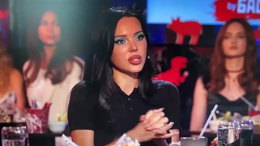 A woman with long dark hair and bright blue eyeshadow is speaking into a microphone. She is wearing a black collared shirt and has her hands clasped in front of her. Behind her, other people are seated at tables in what appears to be a bar or restaurant. The background is dimly lit with neon lights and large screens displaying graphics. To her left, a man with glasses and a grey and black shirt is also seated at a table. The woman is gesturing with her mouth open as if she is in the middle of a sentence. There are drinks and food on the table in front of her.