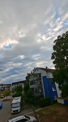 A short video shows a slightly tilted view of a courtyard with buildings and trees under a cloudy sky. In the foreground, the roof of a car is visible. Moving further into the scene, a street with parked cars and a white recreational vehicle is present. Trees with green and yellow leaves are scattered throughout the courtyard. Several multi-story buildings, some with balconies, line the perimeter of the courtyard. The sky is filled with a mix of gray and white clouds, with patches of blue visible in between. The overall impression is a suburban or residential setting on an overcast day.