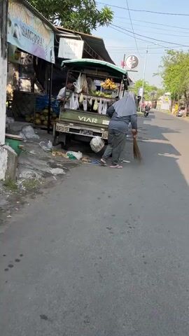 This media file depicts a street scene in Sidoarjo, East Java, likely in the morning or late afternoon due to the long shadows cast.

The primary subject is a person wearing a grey hijab and matching grey clothing, including trousers and a long-sleeved top. They are also wearing pink flip-flops. This individual is standing on the right side of a small, mobile food cart, which is parked on the side of a paved street. The cart is green and appears to be a type of truck or pickup with a stall built on top. It is laden with produce, including bananas hanging in bunches and what appear to be coconuts. Some items are displayed in plastic bags. The cart has the brand name "VIAR" visible on its side, along with a license plate.

Behind the cart, there is a building with a blue facade and awnings. This building appears to be a shop or food establishment, with signage that is partially visible. Text on the building includes "NASI TELUR GULUNG" and "MAKANAN WADE". Further down the street, another building has a sign with "AL-HAMRA" and a circular logo with a silhouette of a figure. A motorcycle is parked in front of this building.

The ground is paved and shows some wet patches, possibly from recent cleaning or rain. There is debris scattered on the ground, including plastic bags and organic waste. The overall atmosphere suggests a typical street scene in a commercial or residential area, with a focus on local commerce. The activity appears to be related to operating or attending to the food cart. The lighting indicates it's daytime, and the weather seems fair.