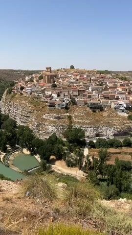 The video shows a panoramic view of a hilltop village under a clear blue sky. The village is characterized by its densely packed buildings with red-tiled roofs, nestled on a rocky incline. A prominent church with a tower stands at the highest point of the village. The terrain surrounding the village is rugged and dry, with some sparse vegetation and trees. In the foreground, a body of water, possibly a river or canal, is visible, with some artificial structures. The overall scene depicts a traditional European village in a somewhat arid landscape.